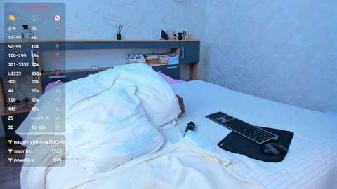 Video of a minimalist bedroom with a white bed, a black keyboard, and a white pillow. The background features a light blue wall and a wooden shelf with a few books and a potted plant.