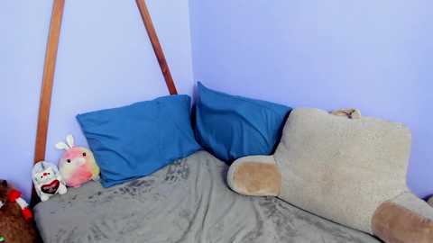 Media: Video of a cozy corner in a room featuring a plush teddy bear and bunny toys on a gray bedspread with blue pillows against lavender walls.