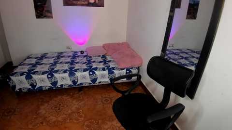 Video of a small, sparsely furnished bedroom with a blue and white patterned bed, pink pillow, and a black chair. A black-framed mirror reflects the room, and purple light creates a soft glow on the white walls.