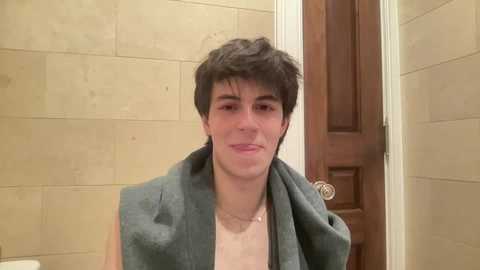 Video of a young man with short, dark hair and a light complexion, wearing a green sleeveless shirt and beige towel. He stands in a beige-tiled bathroom with a wooden door.