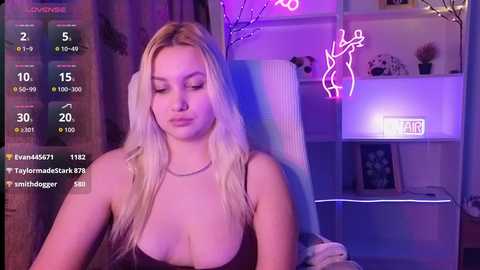 Video of a young blonde woman with long hair, wearing a low-cut top, sitting in a dimly lit room with purple neon lights.