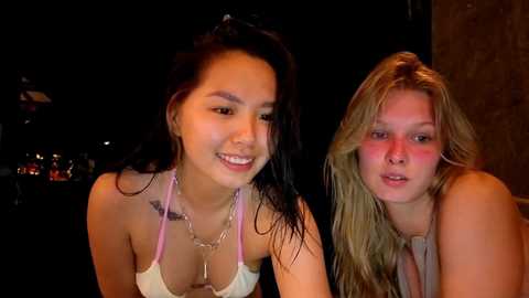 Media: Video of two young women with tanned skin, smiling, in low-light setting. One has long, dark hair, the other blonde. Both wear light-colored tops. Background is dim, possibly a bar.