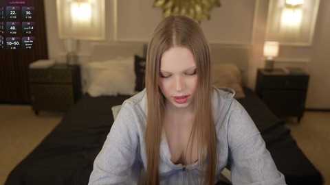 Video of a young Caucasian woman with long straight brown hair, wearing a light grey hoodie, seated on a bed in a modern bedroom with beige walls, dark wooden furniture, and two wall-mounted lamps.