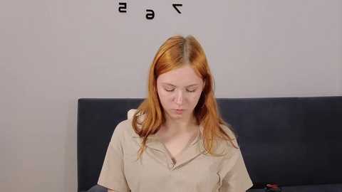 Video of a young red-haired woman in a beige prison jumpsuit, sitting on a dark gray couch against a plain white wall with numbers 23. She appears introspective.