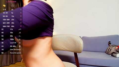 Media: A video shows a woman lifting a purple crop top to reveal her bare back, standing in a living room with a beige couch and a zebra-patterned pillow.