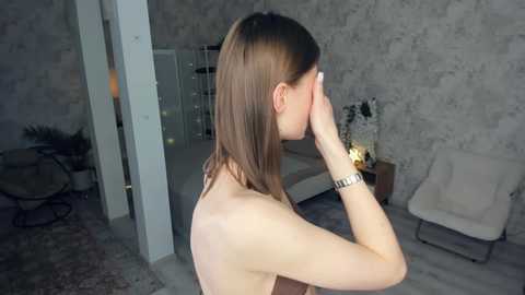 Video of a topless, slender Caucasian woman with long, straight brown hair, wearing a wristwatch, standing with her hand on her forehead, in a dimly lit, modern room with gray walls, a white chair, and a plant.