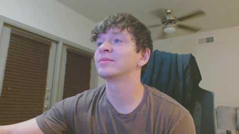 Video of a young man with curly brown hair, wearing a gray t-shirt, sitting indoors. Background features a ceiling fan, closed windows with blinds, and a blue jacket hanging on a chair.