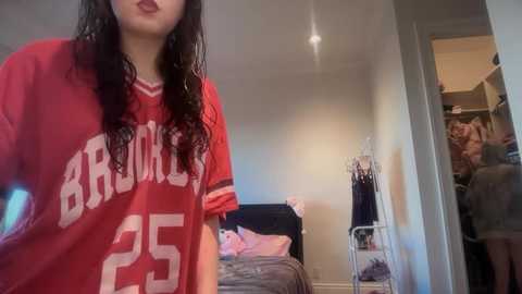 Video of a young woman with long, wet hair wearing a red Rutgers University jersey, standing in a cluttered bedroom with an open closet revealing clothes and a bed.