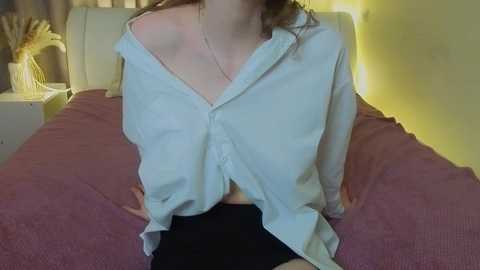 A video of a woman in a light blue button-up shirt, partially unbuttoned, revealing black underwear, sitting on a bed with a pink blanket and a yellow wall.