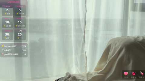 A video of a man with short hair, wearing a beige shirt, lying on a bed with white curtains in the background. The image has a muted color palette.