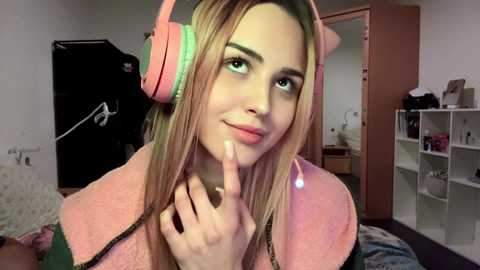 Media: Video of a smiling, light-skinned woman with long blonde hair, wearing pink headphones and a green shirt, indoors with a white shelf and a bed visible.