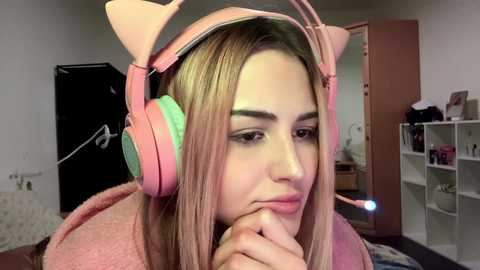 Media: Video of a young woman with long blonde hair, wearing pink cat ear headphones and a fuzzy pink robe, in a cozy bedroom with white shelves and a closed door.
