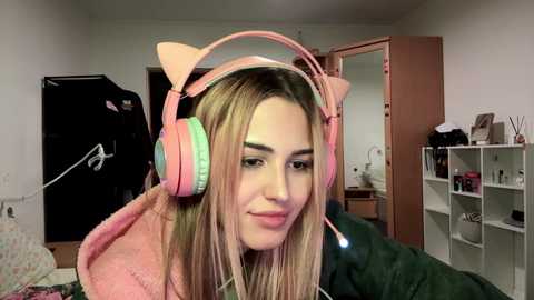 Media: Video of a young woman with straight blonde hair, wearing pink cat ear headphones, smiling in a cozy, cluttered bedroom with a white shelf and a pink door in the background.