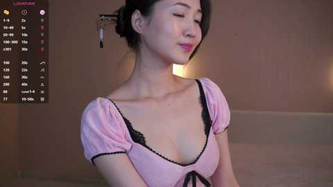 Video of an East Asian woman with fair skin, dark hair in a bun, wearing a pink dress with black lace, smiling softly, set against a beige wall.