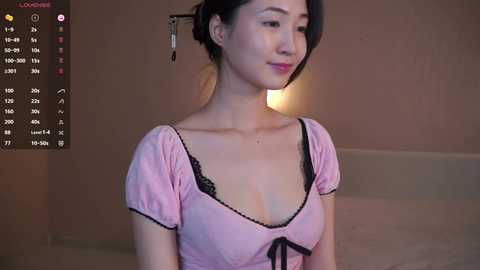 Media: A video of an East Asian woman with light skin, wearing a pink blouse with black lace trim, sitting in a dimly lit room.
