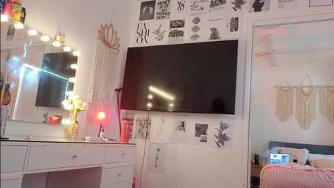 Video of a cozy, girly bedroom with a white vanity table, a large flat-screen TV, a pink macrame wall hanging, and a bed with a pink comforter.