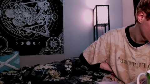 Video of a young man in a beige t-shirt, lying on a bed with a cosmic tapestry and a black floor lamp in a dimly lit room.
