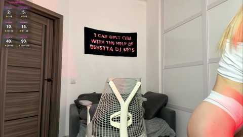 Media: Video of a woman in a white top and pink panties standing in a modern living room with a black banner reading \"I CAN'T CUM WITH THE MOUTH OPEN\" and a grey chair with a checkered blanket.