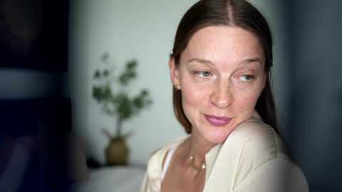 Video of a fair-skinned, blue-eyed woman with straight brown hair, wearing a white jacket, in a softly lit room with a vase of green leaves on a table in the background.