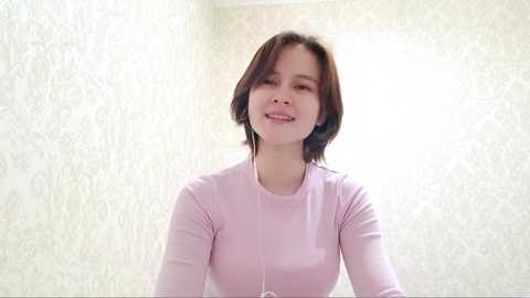 A video of a smiling Asian woman with short, dark hair, wearing a pastel pink long-sleeve top, standing in a room with light-colored, textured wallpaper.