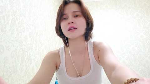 Video of a young woman with short brown hair, wearing a white tank top, looking directly at the camera with a neutral expression. Background features a light, textured wallpaper.