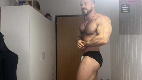 Video of a muscular, bearded man with a dark beard and short hair, wearing black briefs, flexing his biceps, standing in a dimly lit, cluttered room with a white cabinet and a wooden door in the background.