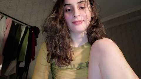 Video of a young woman with curly brown hair, fair skin, and a yellow-green top, smiling softly, in a dimly lit room with hanging clothes and textured wallpaper.