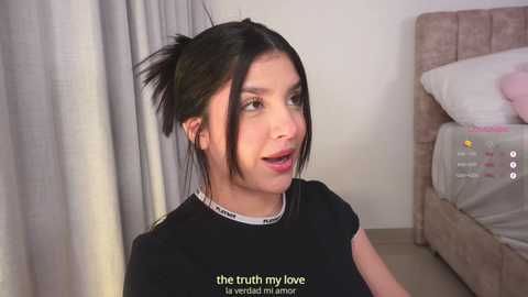 Video of a young woman with dark hair in pigtails, wearing a black t-shirt, sitting in a bedroom with a pink bed and white curtains. Text overlay reads: \"the truth my love.\