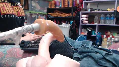 Media: Video of a person lying on a bed with a large, realistic, tan-colored dildo penetrating their mouth, surrounded by a cluttered bedroom with shelves and bottles.