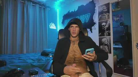 Video of a muscular, shirtless man in a black hat, holding a phone, seated on a gaming chair in a dimly lit, blue-lit bedroom adorned with gaming posters and a bed.