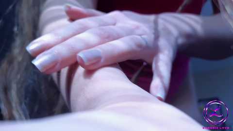 A video of a fair-skinned woman's hand with neatly manicured nails, massaging a bare arm. The background is out of focus, featuring a red fabric. The image has a professional, clinical feel.