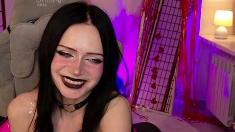 Video of a young woman with pale skin, dark hair, and dark lipstick, smiling, in a dimly lit room with purple and pink lights, a grey chair, and a lamp.