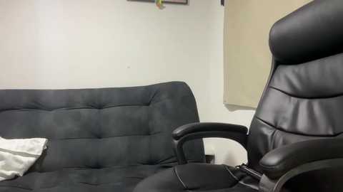 A video of a modern, minimalist room with a black leather gaming chair and a matching black tufted couch against a plain white wall. The scene is simple and clean.