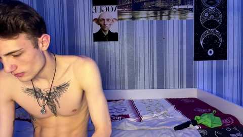 Video of a shirtless young man with short brown hair, wearing a necklace, sitting on a bed with a tattoo on his chest. Blue wallpaper, posters, and a blackboard in the background.