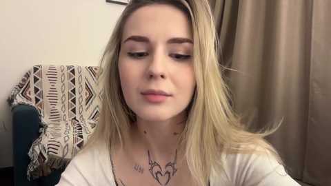 Video of a young woman with fair skin and long, straight blonde hair. She has light makeup, a white top, and a necklace with owl pendant. Background features a patterned blanket and beige curtain.