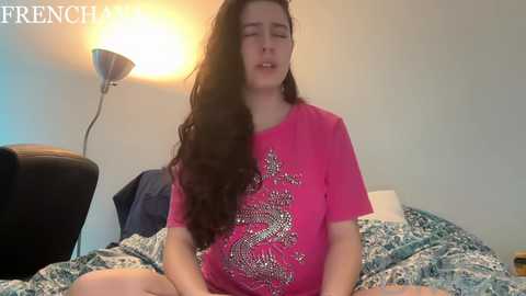 Video of a young woman with long, dark hair, wearing a pink T-shirt with a white floral pattern, sitting cross-legged on a bed with a patterned duvet. The room has a white wall, black leather chair, and a lamp emitting warm light.