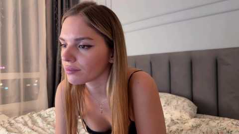 Media: Video of a light-skinned, slim young woman with long, straight, blonde hair, wearing a black tank top, sitting on a bed with floral-patterned bedding in a modern bedroom.