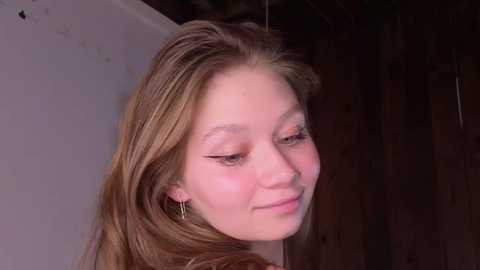 Video of a young Caucasian woman with light skin and long, straight blonde hair. She has a gentle smile, closed eyes, and wears gold hoop earrings. Background is dark wooden paneling and a white wall.