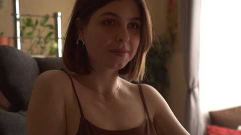 Media: Video of a young woman with shoulder-length brown hair, wearing a brown tank top, sitting indoors. She has a neutral expression. Background includes a dark sofa, green curtains, and a plant.