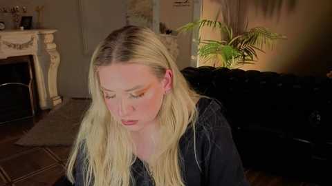 Media: Video of a fair-skinned, blonde woman with long hair, wearing a black jacket, sitting on a black leather couch, surrounded by a cozy living room with a fireplace, fern, and warm lighting.