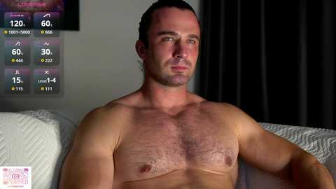 Video of a shirtless, muscular white man with short dark hair, sitting on a white couch. Background includes a dark curtain and a digital fitness app screen displaying stats.
