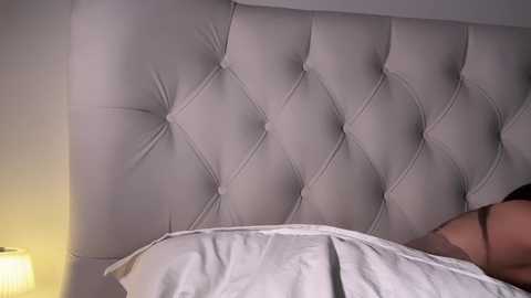 Media: Video of a modern, plush, tufted headboard in a neutral color, with a white bedsheet partially covering a person's upper torso and arm, suggesting a cozy bedroom setting.
