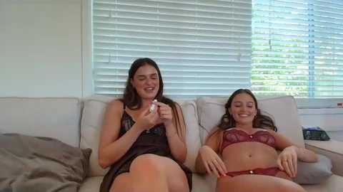 A video of two young women, one in a black lace dress, the other in a maroon lingerie set, sitting on a beige sofa, smiling, in a bright room with white blinds and a blue sky view.