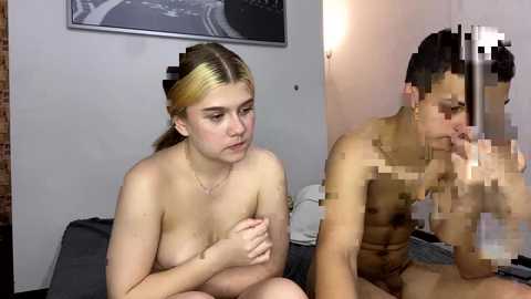 Video of a nude, fair-skinned young woman with blonde hair, covering her breasts, and a nude, darker-skinned man with a penis, both in a bedroom setting.
