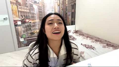 Video of an Asian woman with long black hair, wearing a striped kimono, sitting on a bed with European cityscape wallpaper in the background.
