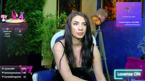 Video of a young woman with long black hair, wearing a black dress, sitting in front of a blue backdrop with a plant and a microphone. Live streaming on \"Lovesense ON.\