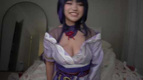 A video of a smiling East Asian woman with dark purple hair, wearing a revealing, purple, lace-trimmed cosplay outfit with a deep cleavage, standing in a dimly-lit bedroom.