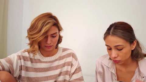 Video of two women with light skin, one with short, wavy, blonde hair, the other with straight, dark brown hair, both wearing casual striped sweaters, sitting close, looking down, indoors with a neutral background.