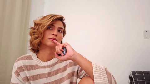 Video of a young, light-skinned woman with wavy, shoulder-length blonde hair, wearing a beige and white striped sweater, holding a finger to her lips in a thoughtful pose, in a minimalist room with white walls and a gray plaid blanket in the background.