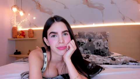 Video of a Latina woman with long dark hair, fair skin, and full lips, lying on a bed with floral pillows, wearing a black tank top, in a cozy, dimly lit room with beige walls and modern decor.